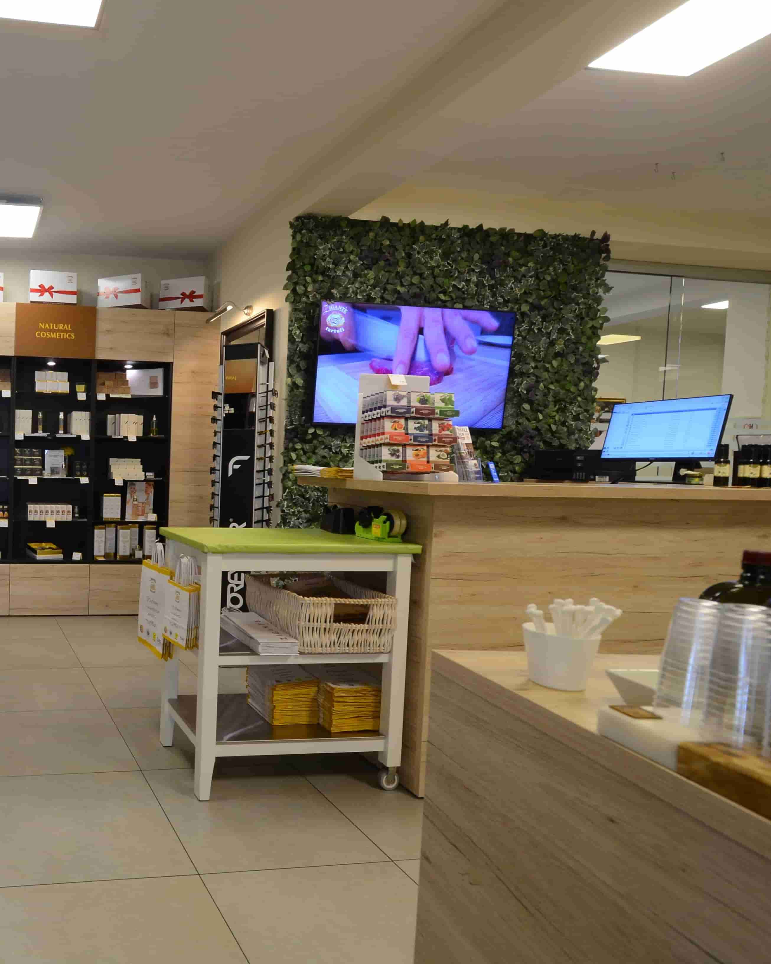 A retail store interior with a wooden counter, a green leafy wall backdrop, a mounted TV showing a close-up, and a small white cart with a green top in the foreground.