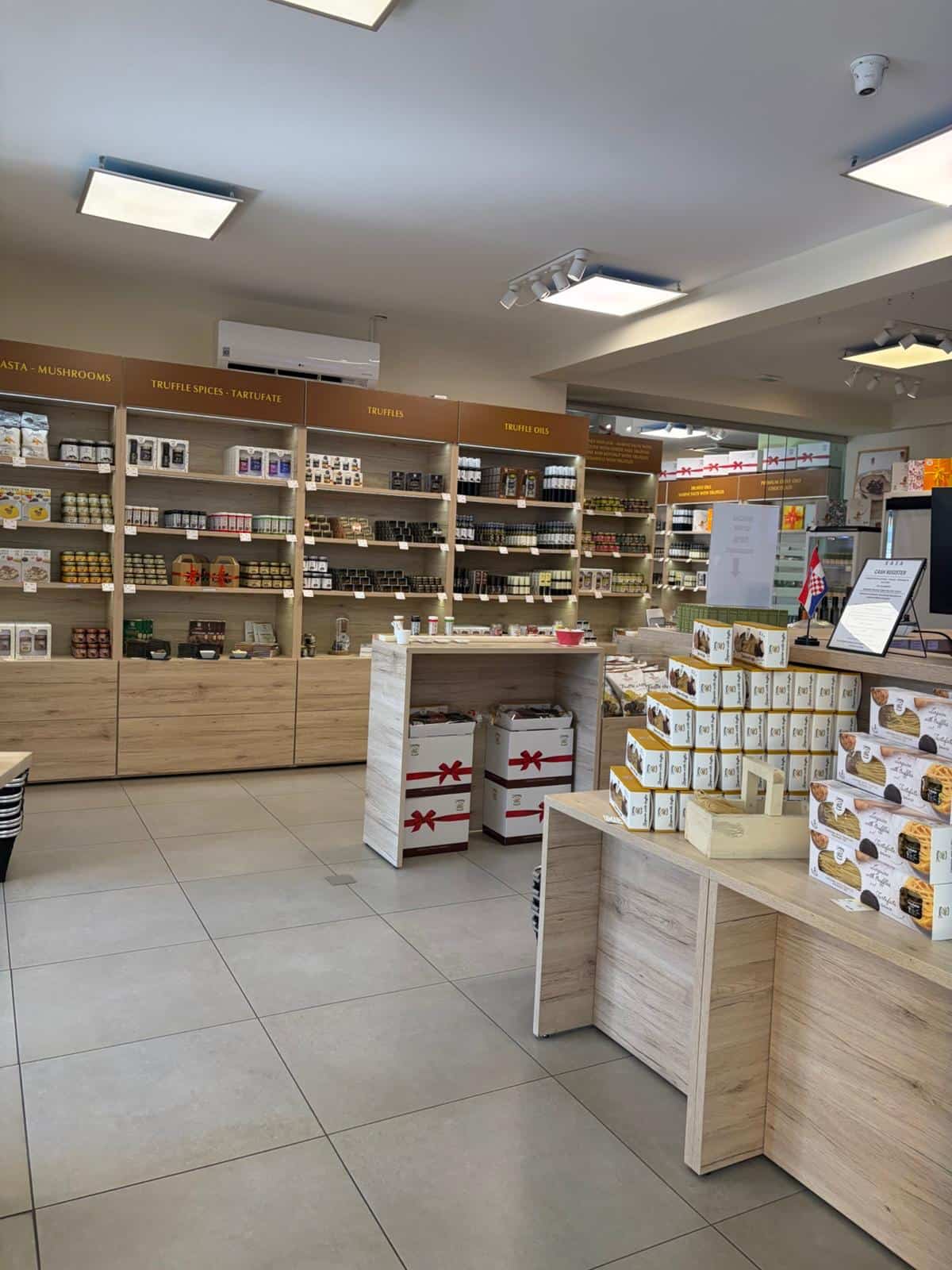 A retail store interior with light wood shelving stocked with packaged goods, a tiled floor, overhead ceiling lights, and a counter area on the right side.