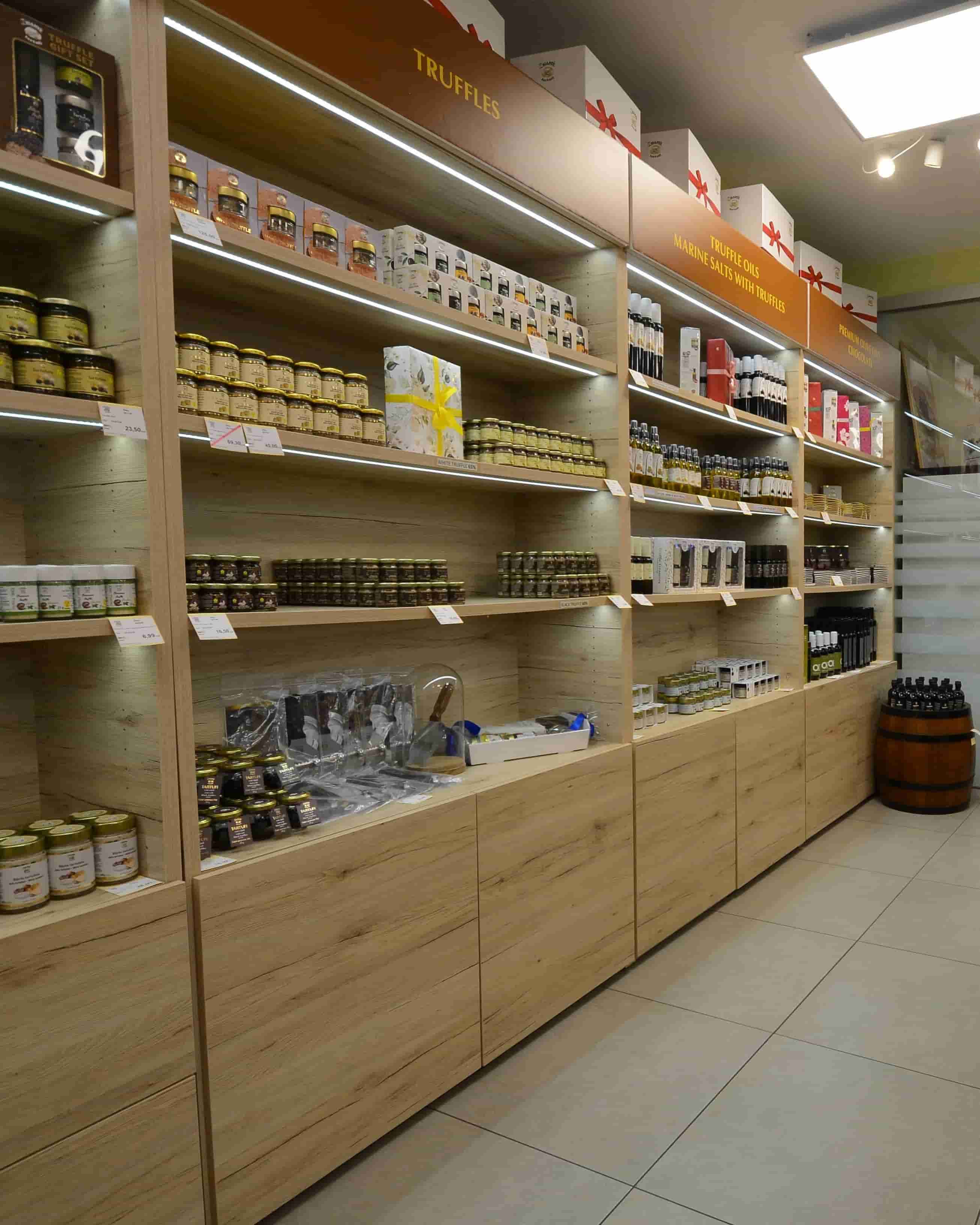 Wooden retail shelving units stocked with assorted jars, boxes, and packaged goods in a store interior. Signs above shelves include the word TRUFFLES, and a barrel-style bucket sits on the floor to the right.