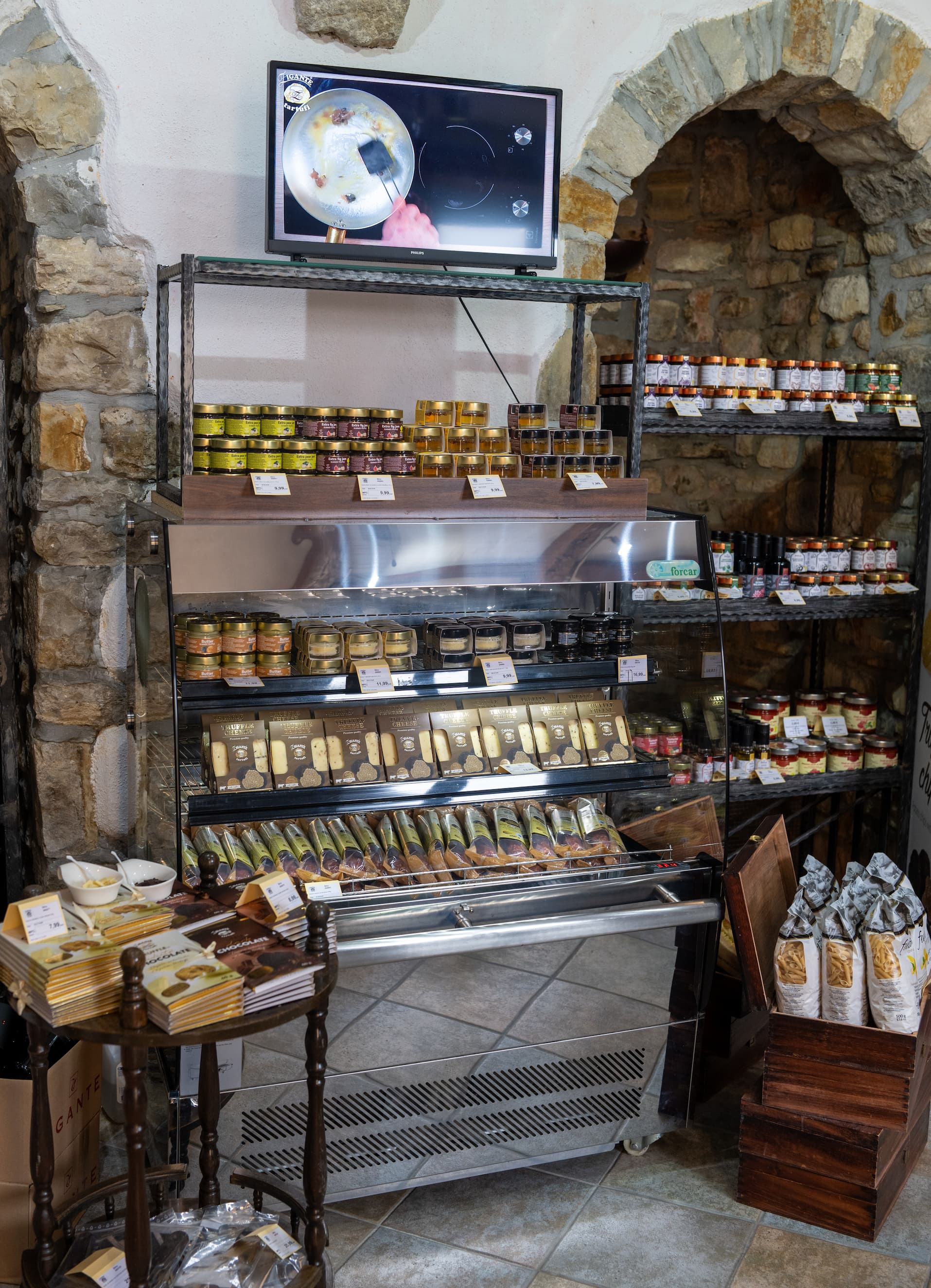 A glass-front refrigerated display case filled with truffle salami, truffle  cheeses and butter with truffles. A set in a rustic stone-walled shop with shelves of packaged food gourmet products and a small TV mounted above.