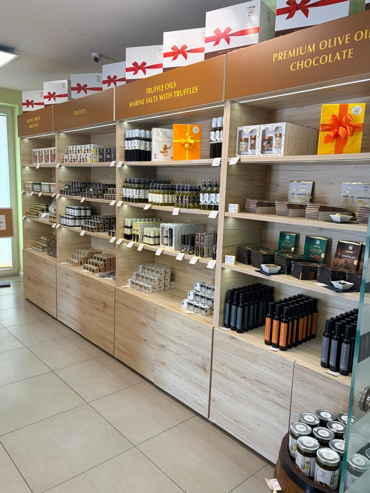 A retail display shelf filled with packaged food products, including bottles, boxes, and jars, under warm lighting. Visible signage includes 'PREMIUM OLIVE OIL, CHOCOLATE' and 'TRUFFLES OIL'.