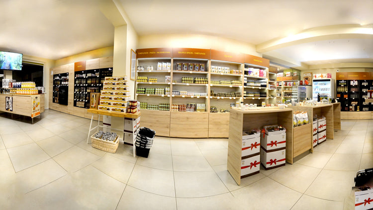 A wide interior view of a retail store or supermarket aisle with wooden display counters, shelves stocked with packaged goods, tiled floors, and bright overhead lighting.